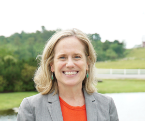 Kristen Miale smiles outside in front of field and pond with white fence.