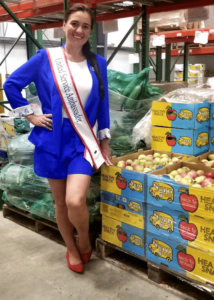 Misty Coolidge, United Service Ambassador for Worldwide USA Pageants in Good Shepherd Food Bank's Auburn distribution center. She's standing in front of produce in a blue suit and wearing her sash.