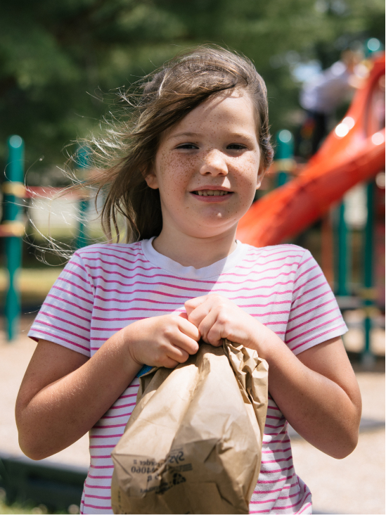 A child holding a bagged lunch and smiling on the playground. 