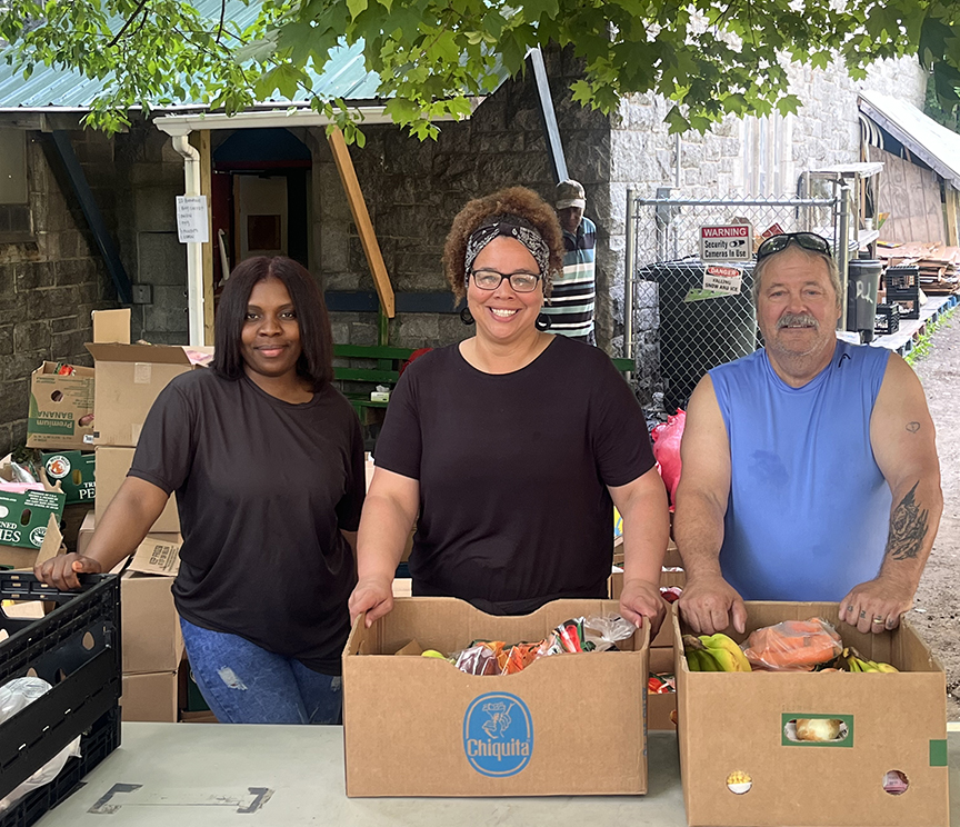 Two women in black shirts and a man in a blue shirt pose with food boxes at a pantry distribution