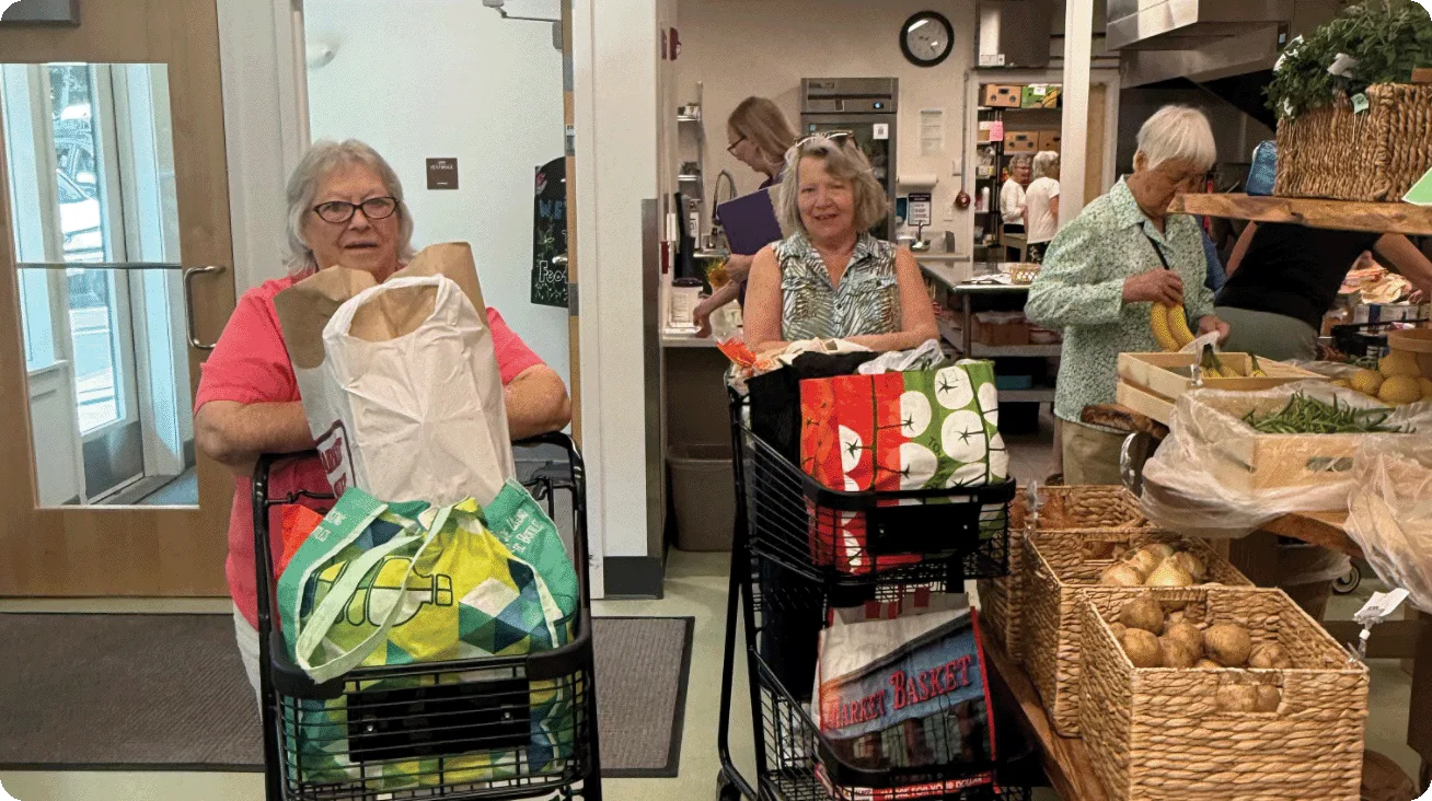 Carol (left) stands behind a cart filled with food inside Footprints Food Pantry, a partner of Good Shepherd Food Bank.