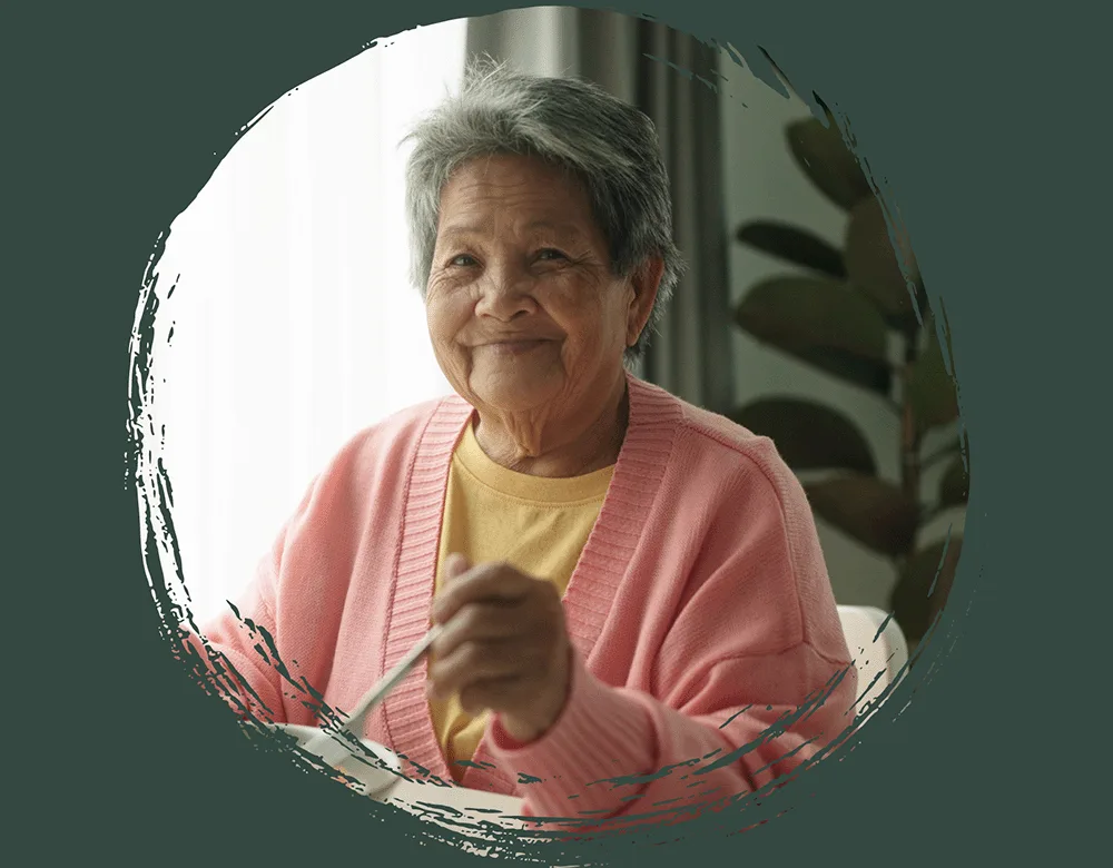 Give Now Smiling elderly woman with short gray hair wearing a pink cardigan and yellow shirt, sitting indoors near a window, holding a utensil.
