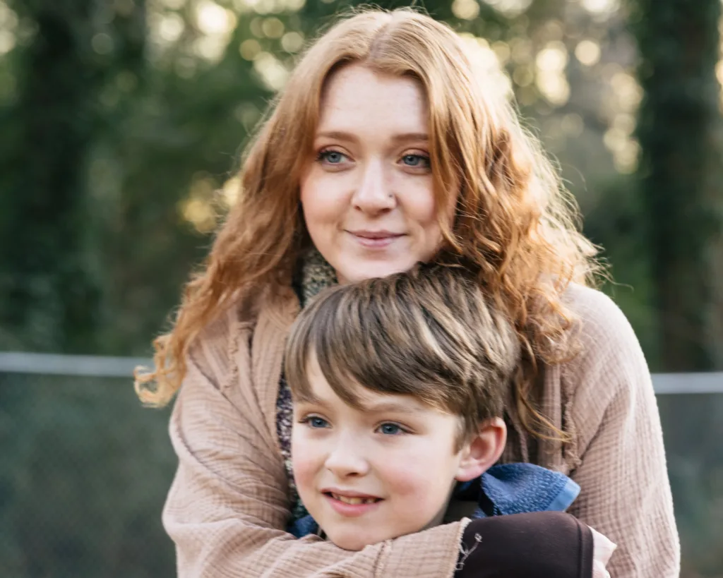 A woman with long red hair hugs a young boy from behind outdoors, both smiling and looking ahead with trees blurred in the background.