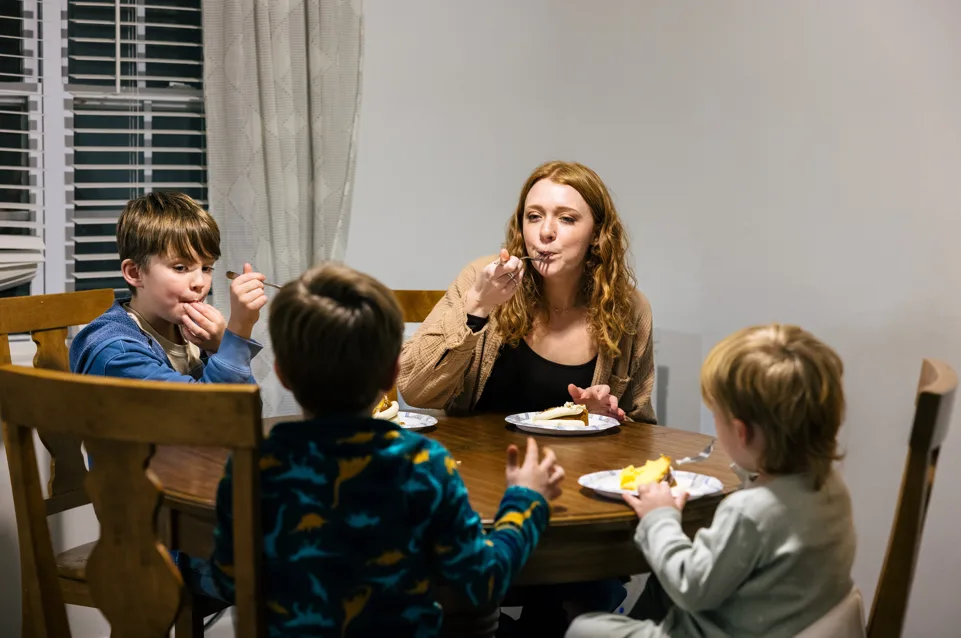 A woman and three children sit around a wooden table, eating together in a cozy, well-lit room.