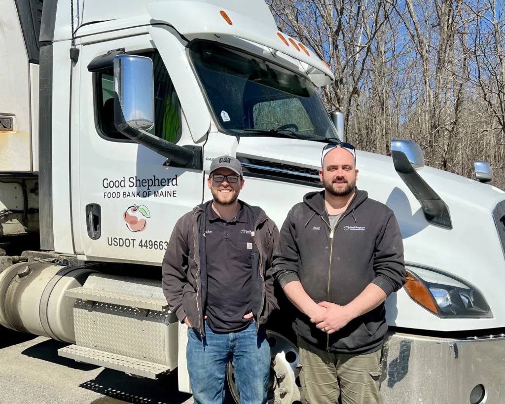 Two men in work clothes stand smiling in front of a Good Shepherd Food Bank of Maine truck on a sunny day with leafless trees in the background.