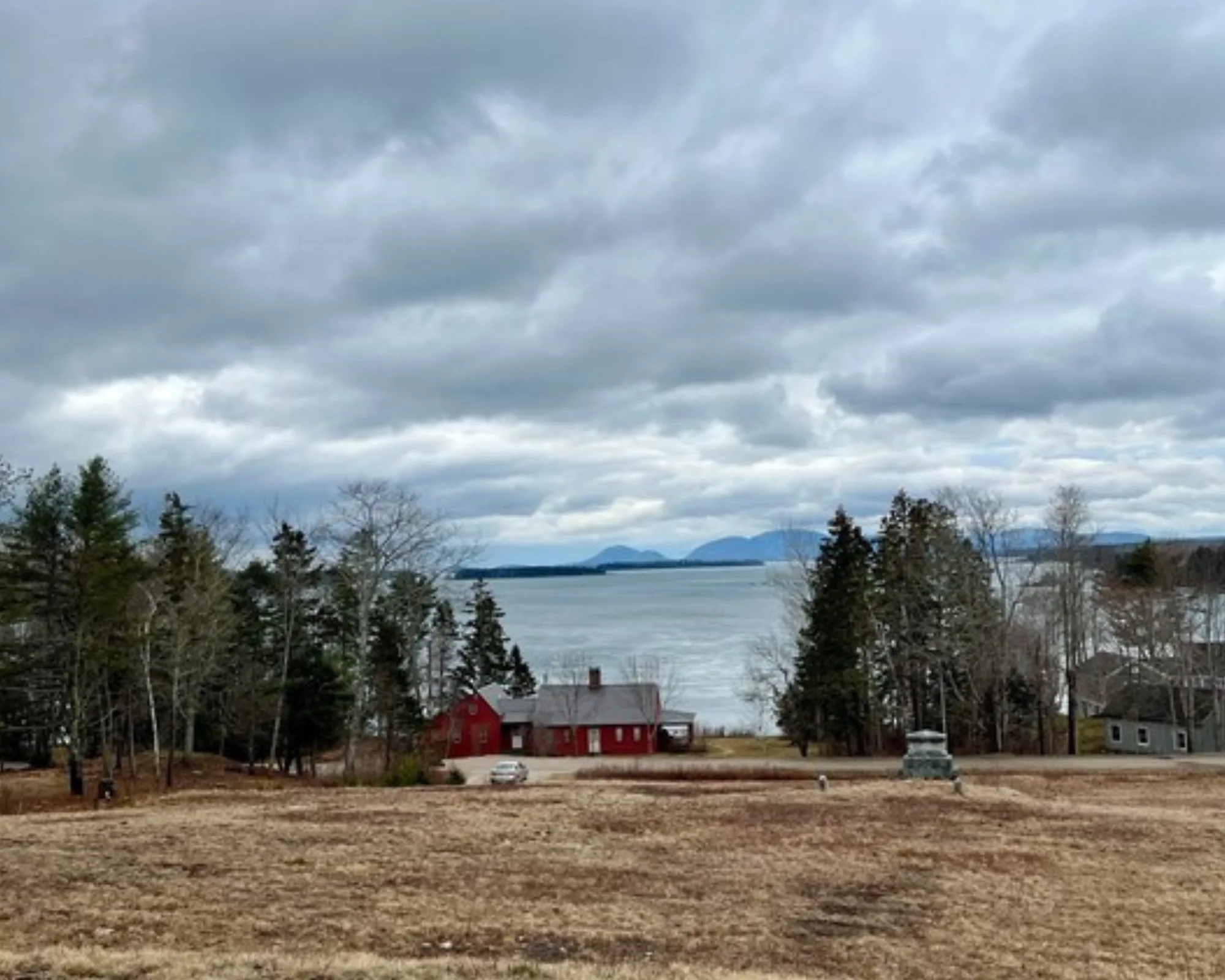 A cloudy sky over a lake with mountains in the distance, trees by the shore, and a red house and monument in a grassy, brown field.