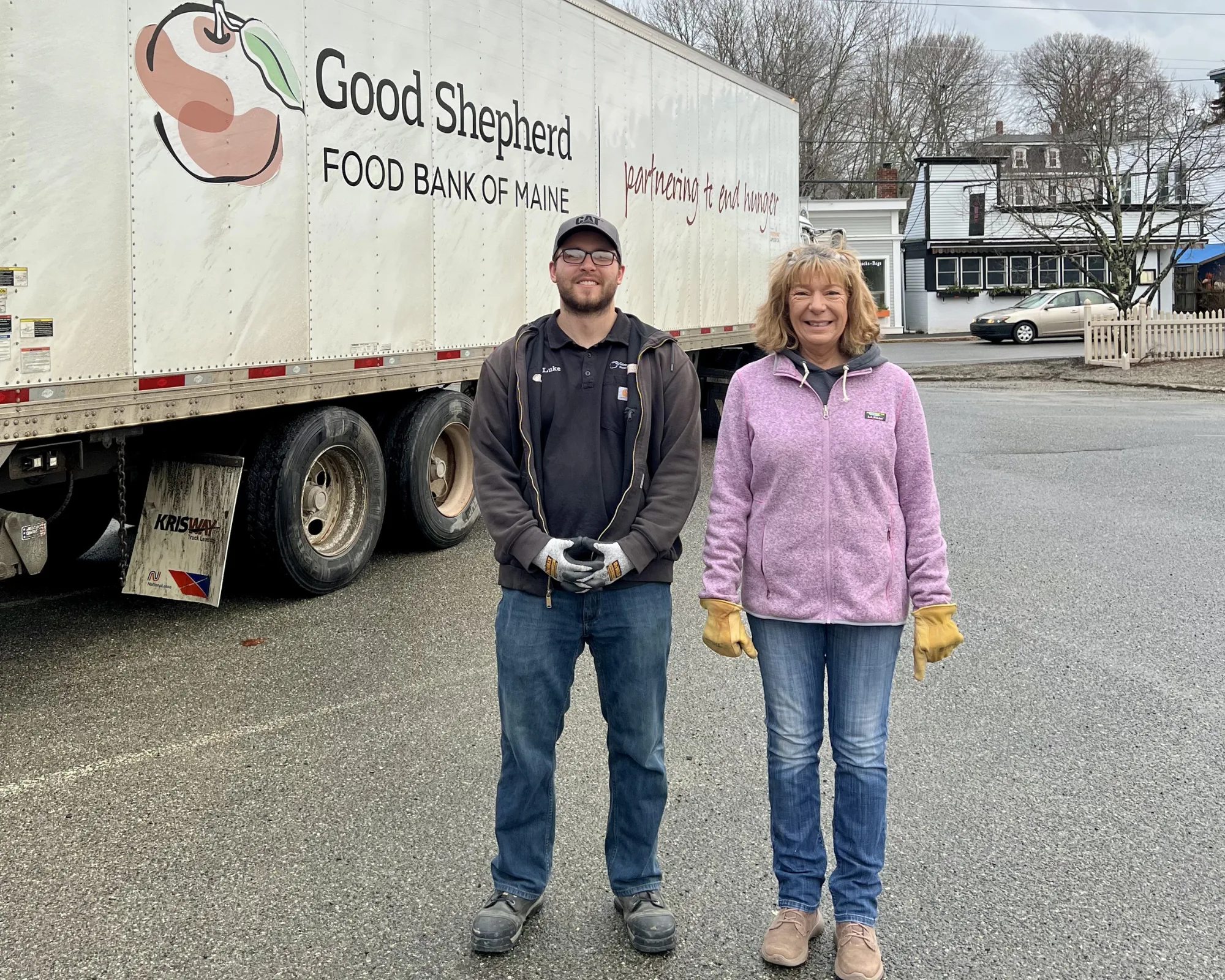 Two people stand smiling in front of a Good Shepherd Food Bank of Maine truck in a parking lot on a cloudy day.