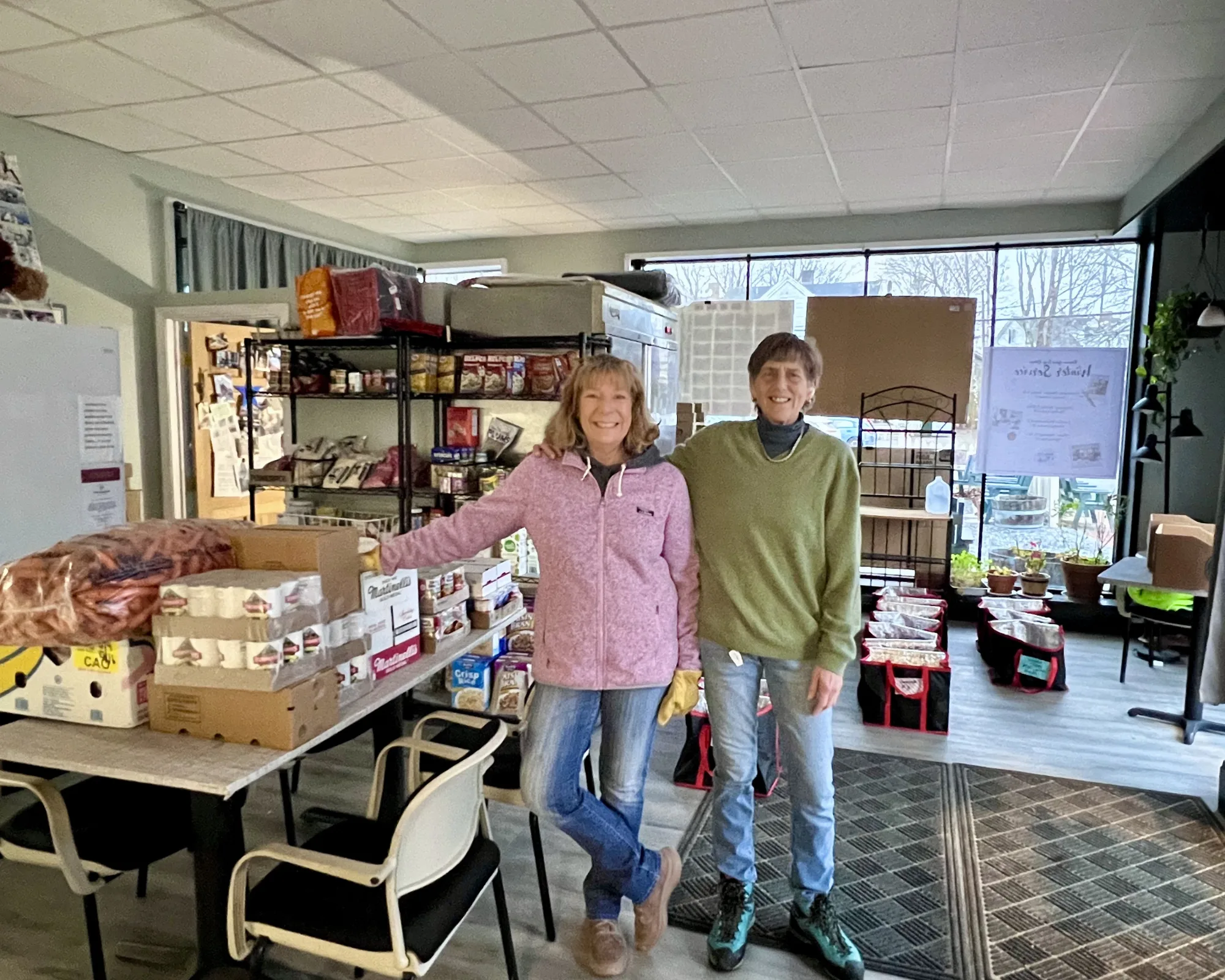 Two women stand smiling in a room with shelves of food supplies, boxes, and pet products. The setting looks like a community or donation center.