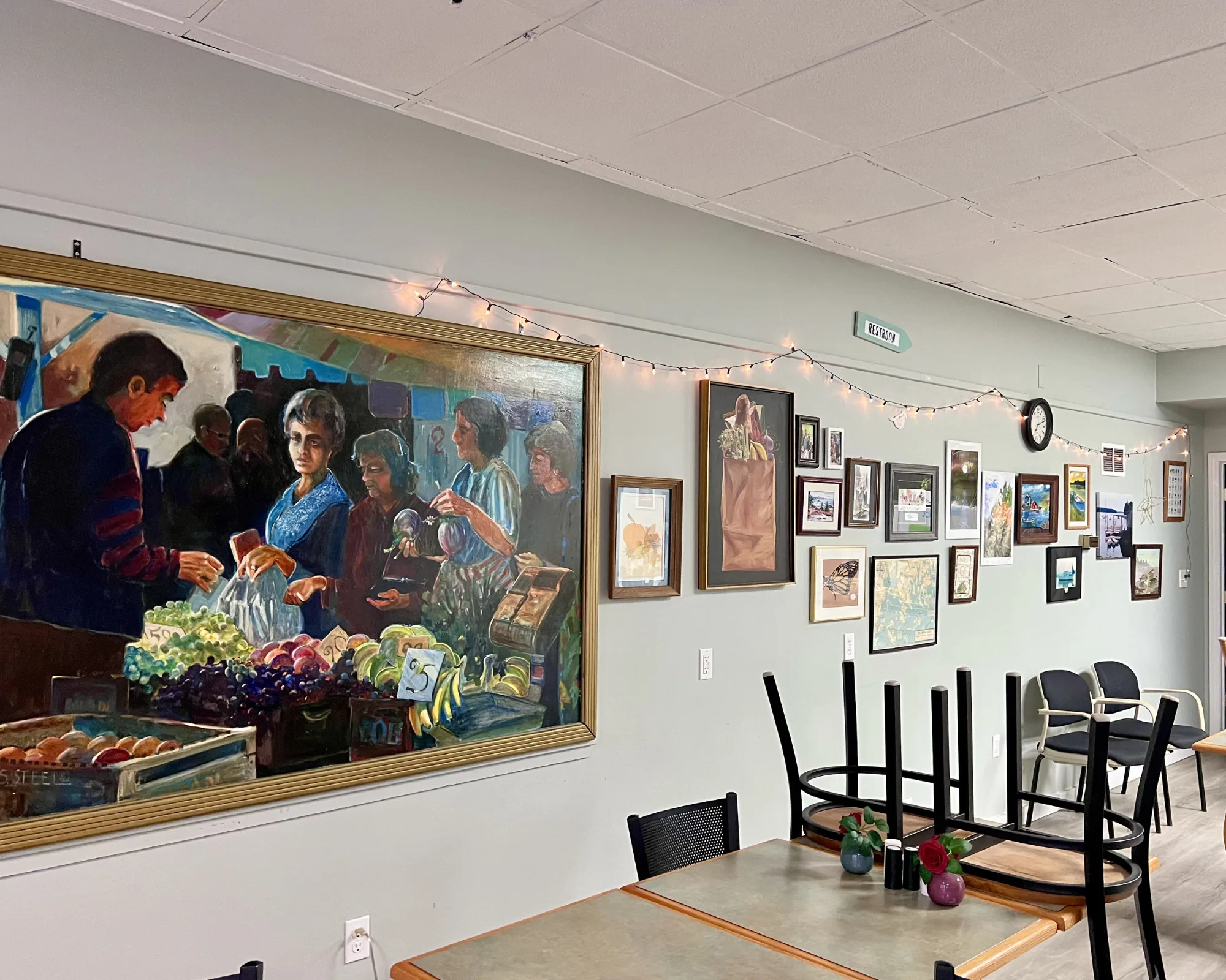 A cozy café wall decorated with framed art, fairy lights, and a large painting of a market scene; chairs are stacked on tables in the foreground.