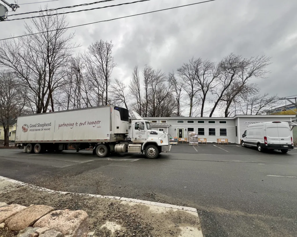 A Good Shepherd Food Bank truck parked outside a building on a cloudy day, with leafless trees and a white van nearby.