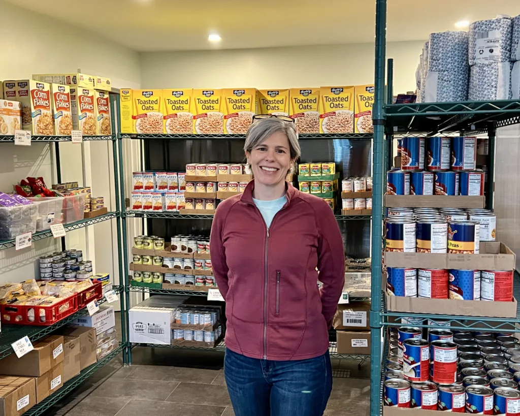 A woman in a red jacket stands smiling in a food pantry, surrounded by shelves stocked with cereal boxes, canned goods, and other food items.