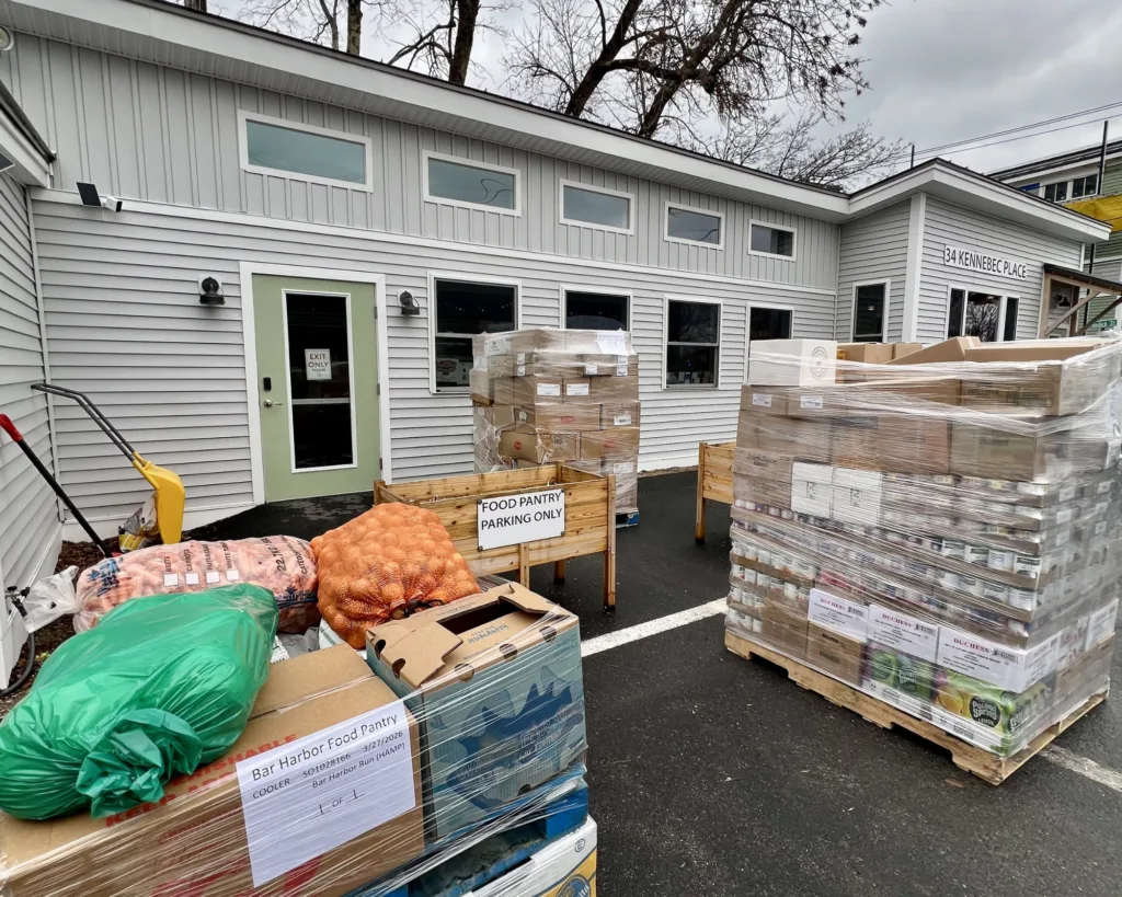 Boxes and pallets of food are stacked outside a food pantry building, with signs indicating it is for food distribution.