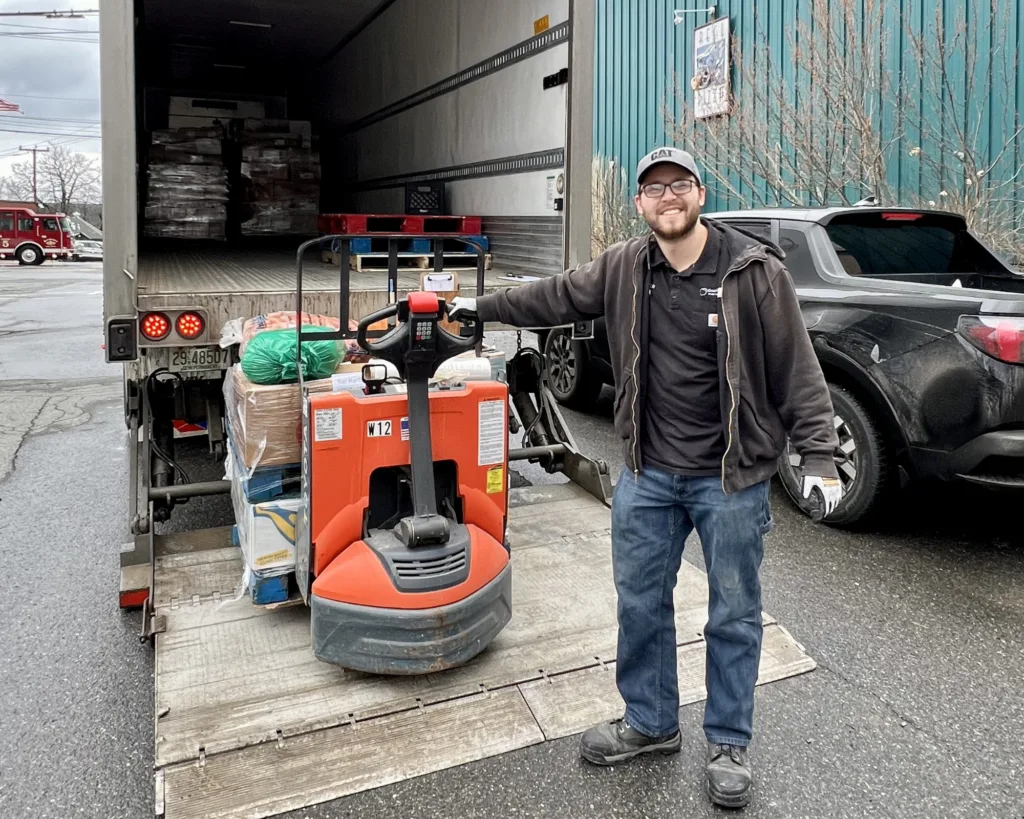 A smiling man stands next to a pallet jack loaded with goods on a truck lift gate, ready for unloading on a wet street.