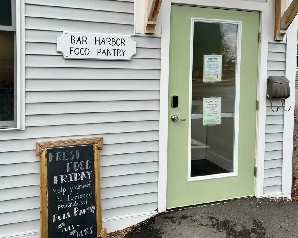 Entrance to Bar Harbor Food Pantry with a green door. Signs display hours, donation info, and a chalkboard advertises Fresh Food Friday for leftover perishables.