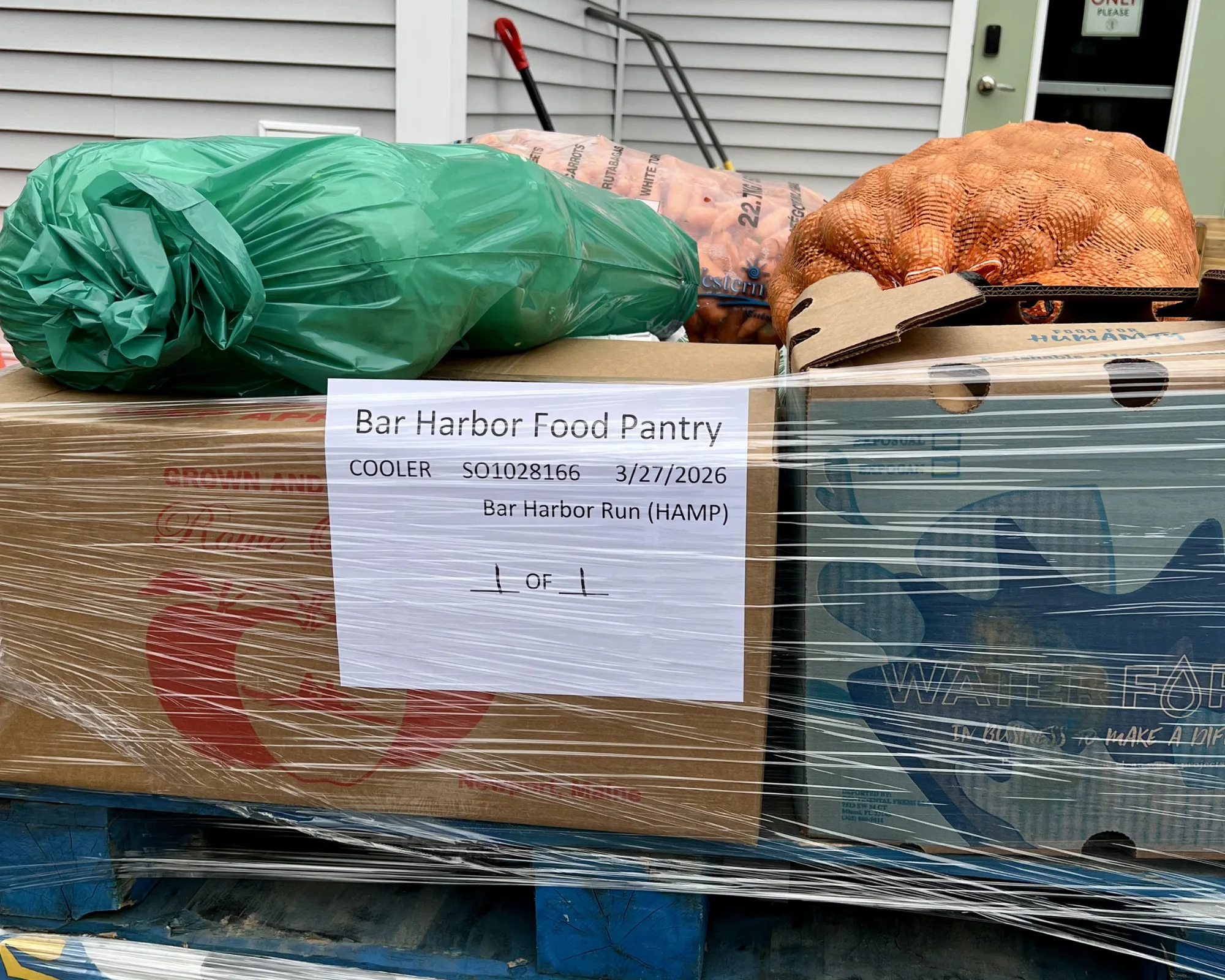 A pallet with boxes of food wrapped in plastic, labeled Bar Harbor Food Pantry with a date of 3/27/2026. A green bag and a mesh bag of onions are on top.