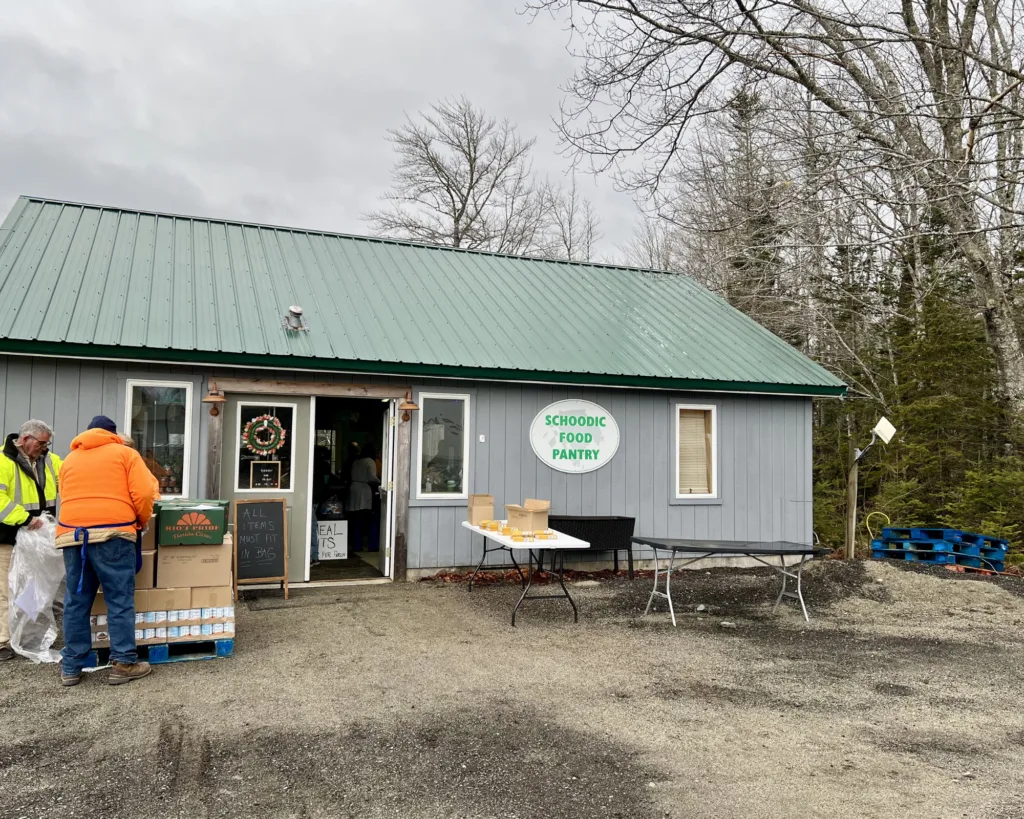 A small gray building with a green roof labeled Schoodic Food Pantry; people stand outside next to boxes and tables on a cloudy day.