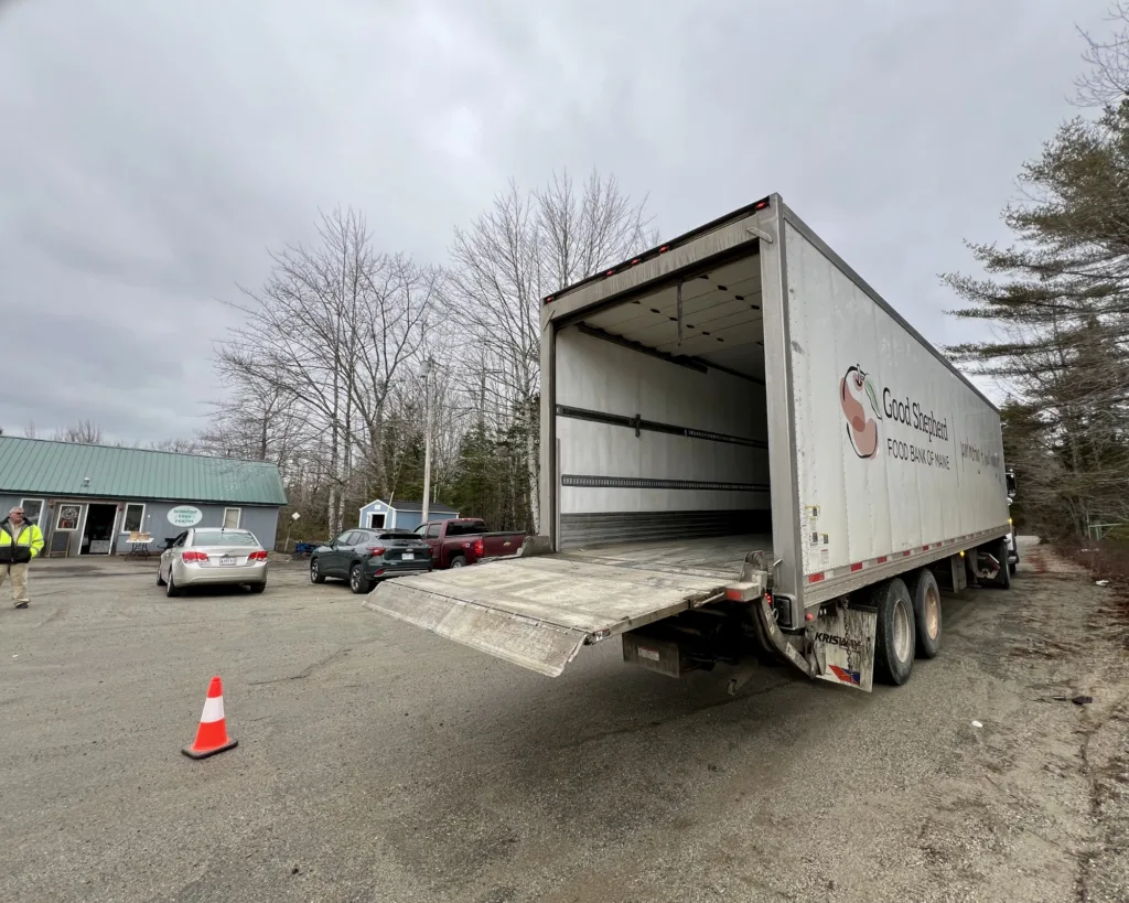 A large delivery truck with its ramp down is parked on a lot near several cars, a building, and orange traffic cones on a cloudy day.