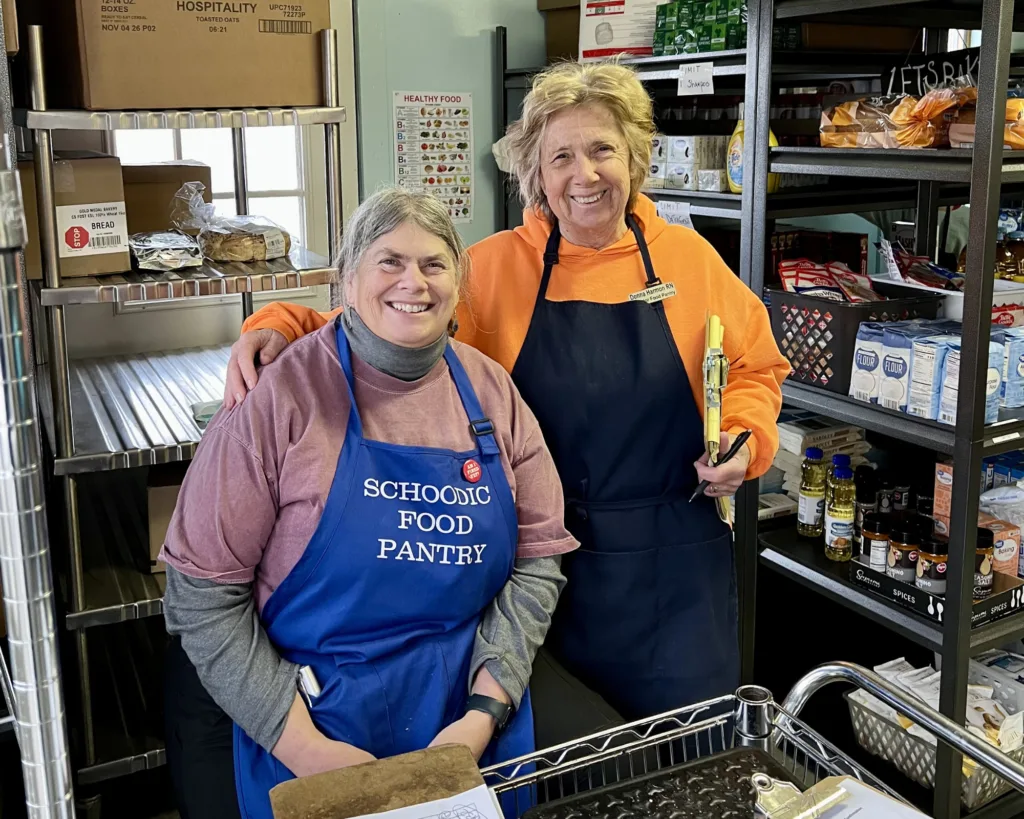 Two smiling women wearing aprons stand in a food pantry surrounded by shelves stocked with food and supplies. One holds a clipboard; the other holds a notepad and pen.