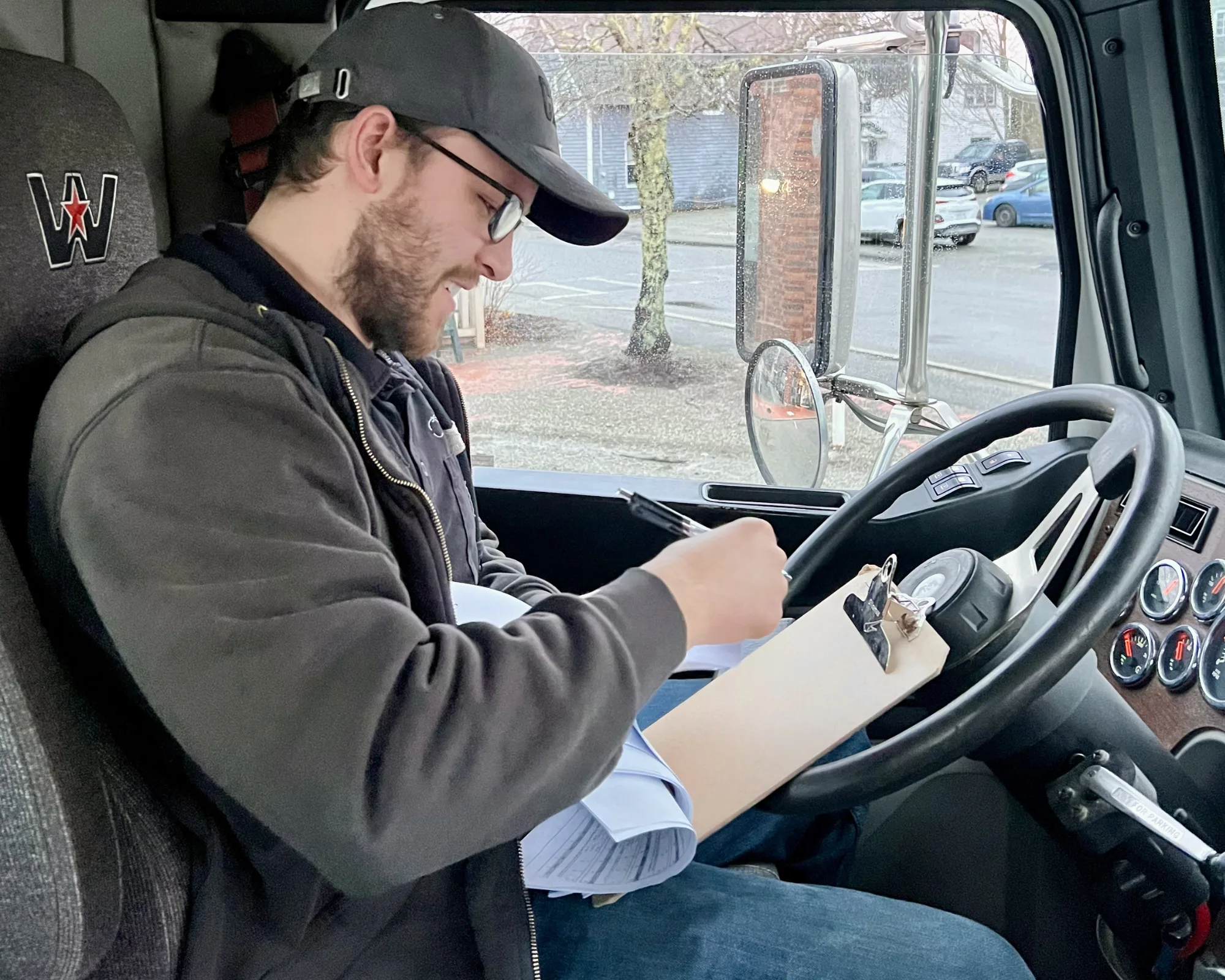 A man wearing glasses and a cap sits in a truck cab, writing on a clipboard with a pen.