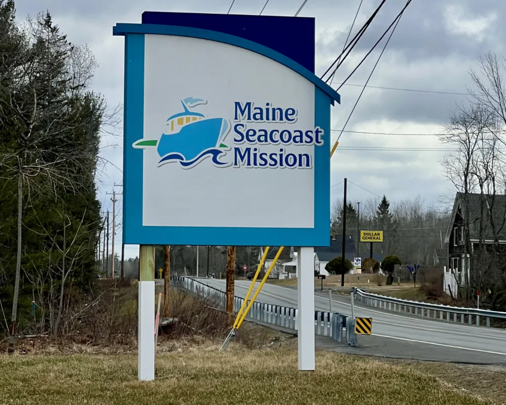 A roadside sign reads Maine Seacoast Mission with a boat graphic above it. Trees, power lines, and a road with guardrails are in the background under a cloudy sky.