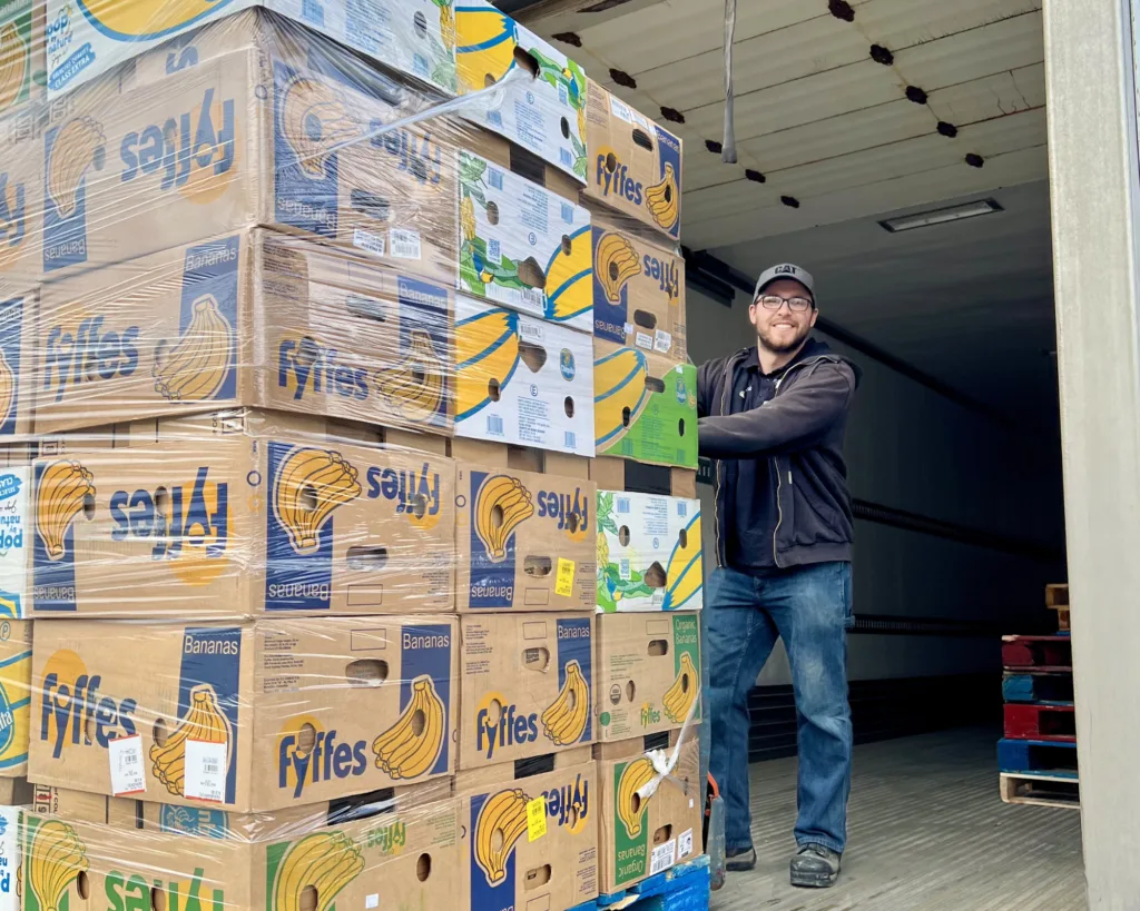 A smiling person stands next to a large stack of Fyffes banana boxes inside a truck trailer, ready for unloading or delivery.