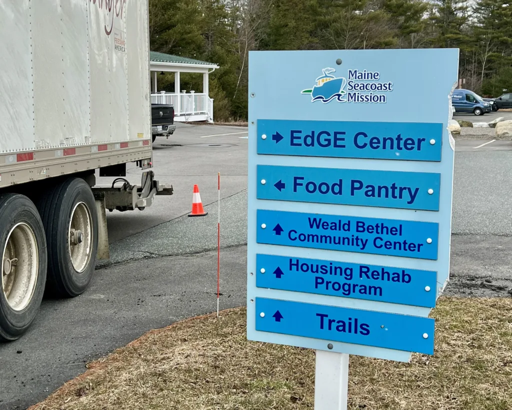 A blue directional sign for Maine Seacoast Mission points to EdGE Center, Food Pantry, Weald Bethel Community Center, Housing Rehab Program, and Trails. A truck is parked nearby.