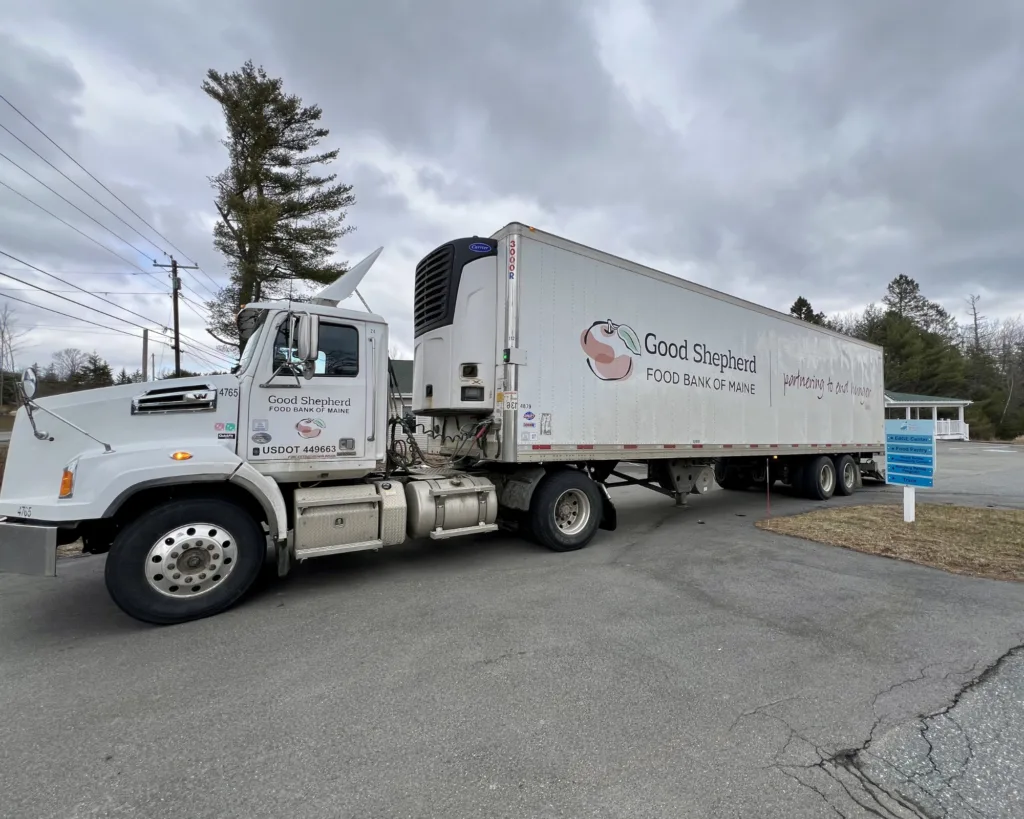 A white Good Shepherd Food Bank of Maine truck is parked on a paved road under a cloudy sky, near trees and a small building.