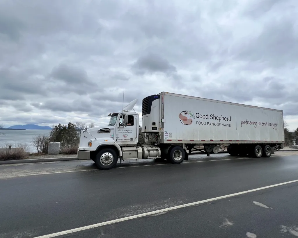 A Good Shepherd Food Bank of Maine truck is parked on a roadside with trees, water, and mountains in the background under a cloudy sky.