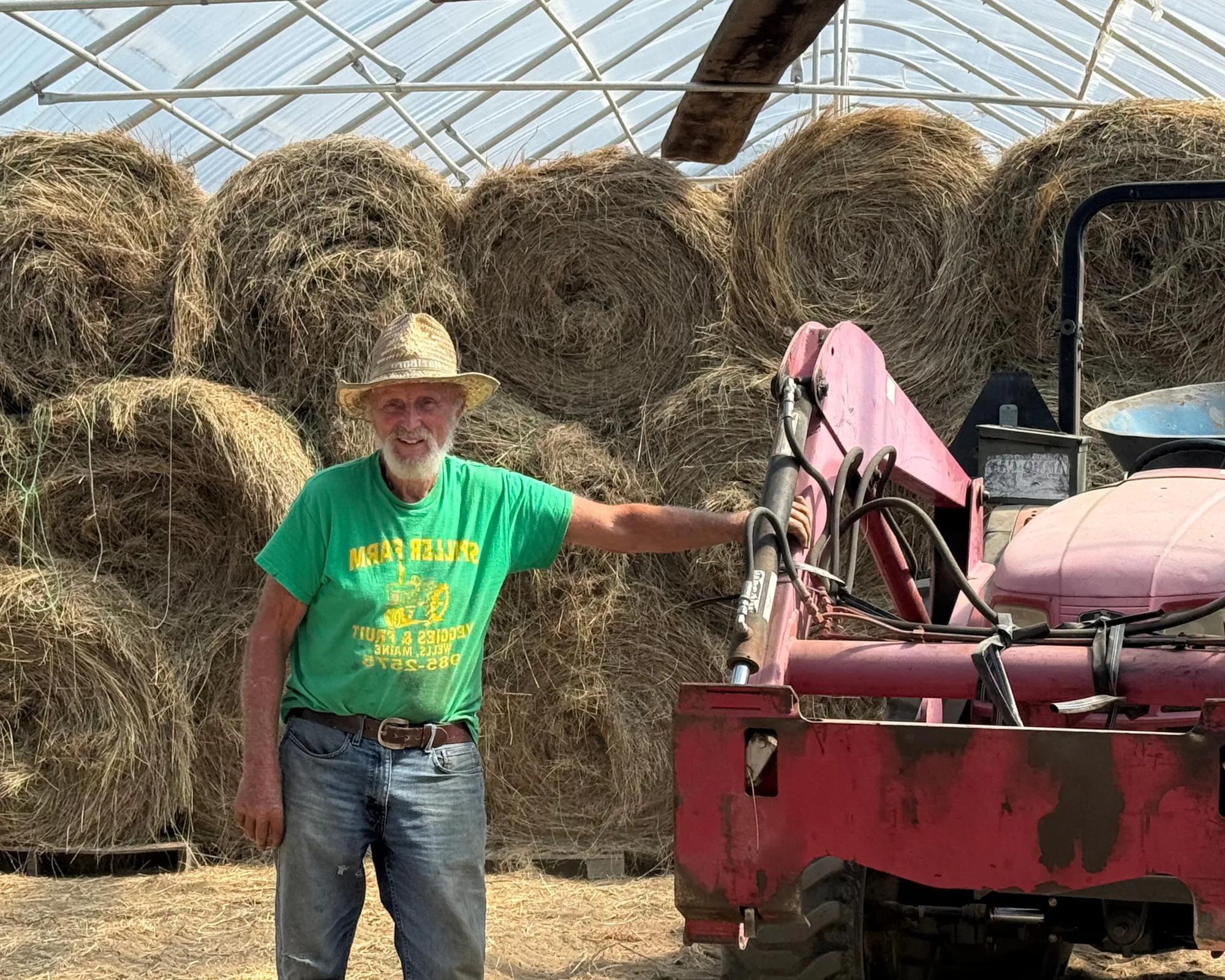 An older man in a straw hat and green t-shirt stands smiling beside a red tractor, with large hay bales stacked behind him in a greenhouse.