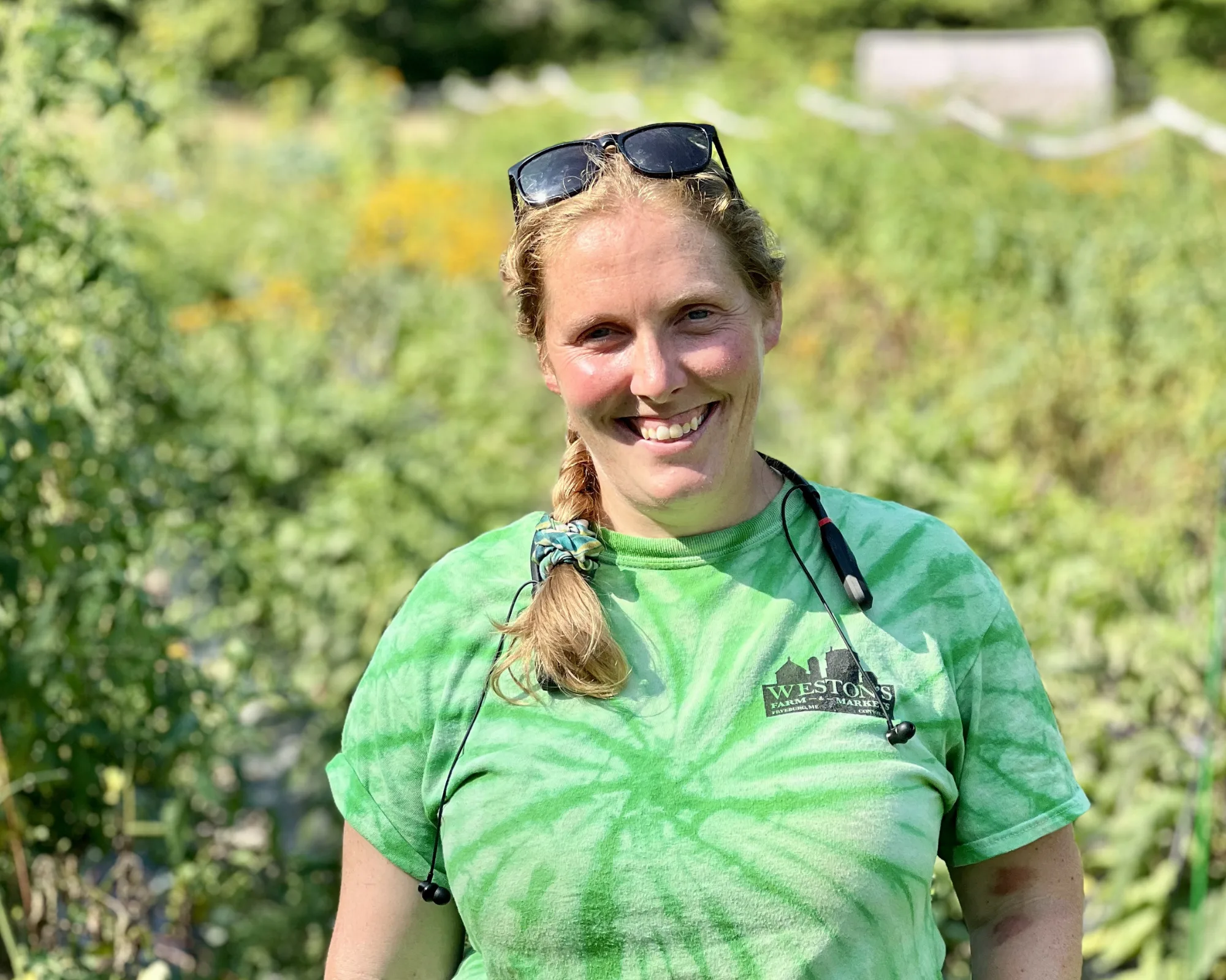 A smiling woman in a green tie-dye shirt stands in a sunny garden, wearing sunglasses on her head and a cord around her neck.