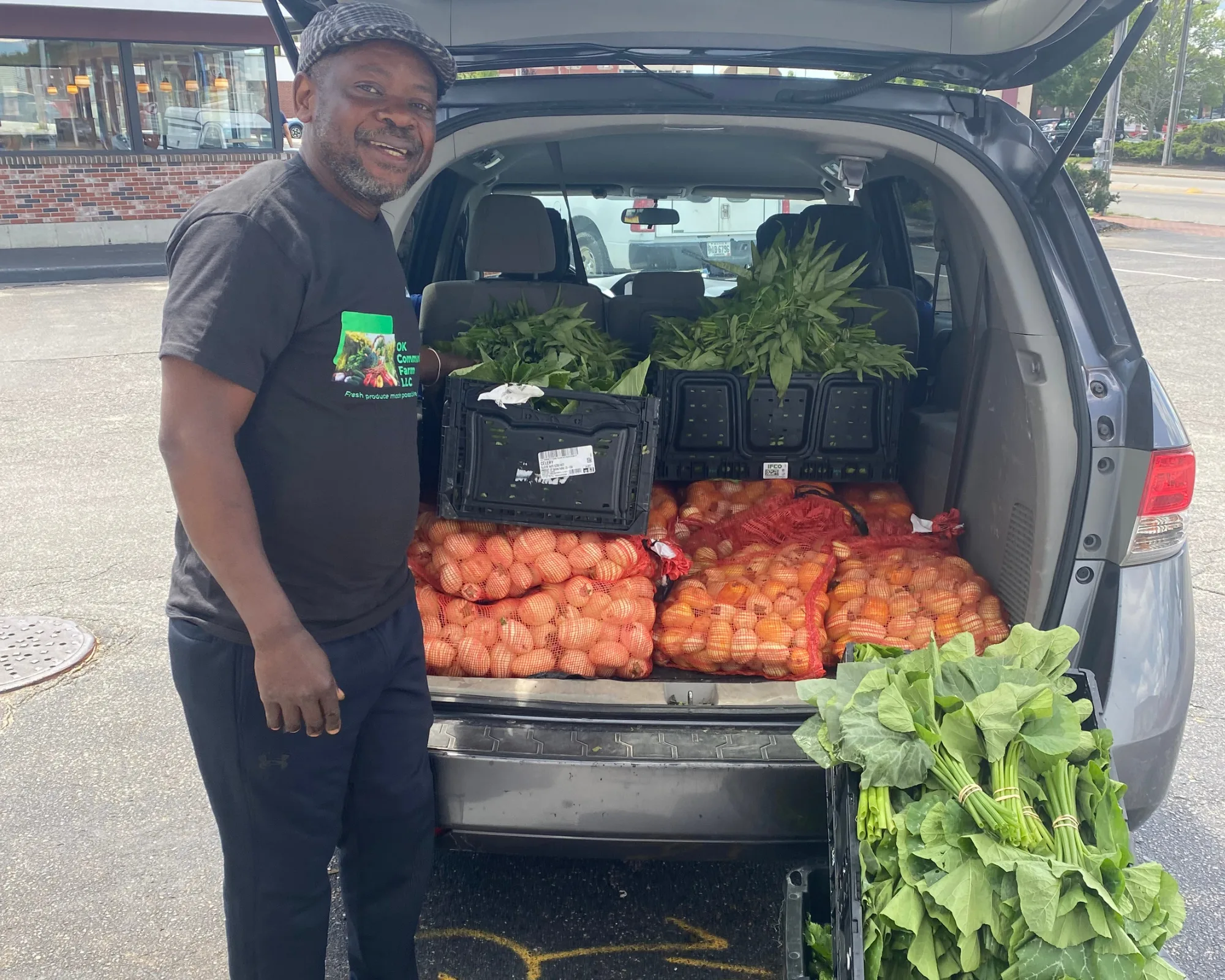 A man smiles next to an open car trunk filled with bags of onions, leafy greens, and crates of fresh vegetables in a parking lot.