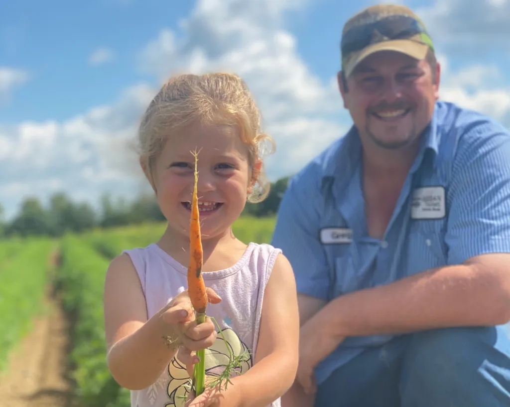 Smiling young girl holding a freshly picked carrot in a field, with an adult man kneeling and smiling in the background under a blue sky.