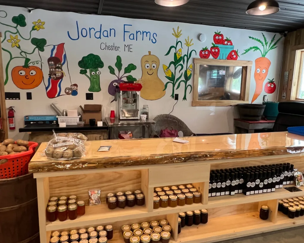 A farm store counter with jars of preserves and bottles, in front of a colorful mural of smiling vegetables labeled Jordan Farms, Chester, ME.