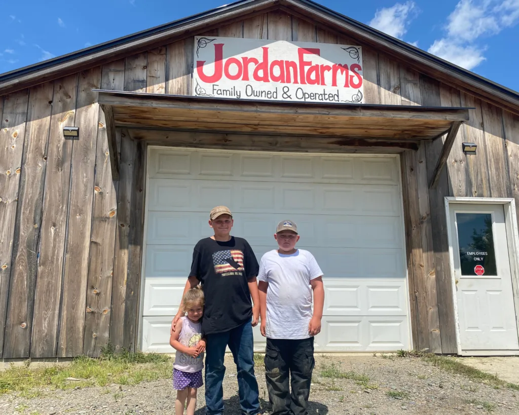 Three children stand in front of a wooden building with a sign that reads Jordan Farms, Family Owned & Operated on a sunny day.