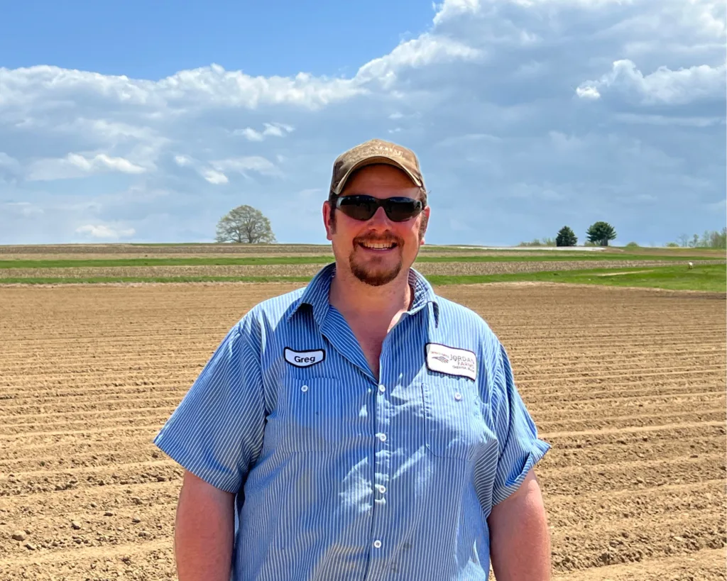A man wearing sunglasses, a cap, and a striped shirt stands smiling in front of a freshly plowed field under a partly cloudy sky.