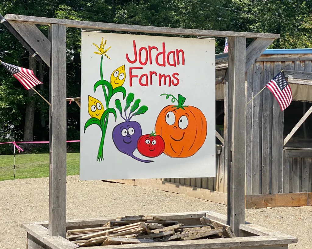 A sign for Jordan Farms features cartoon vegetables and a pumpkin with smiling faces, surrounded by small American flags on a wooden frame.