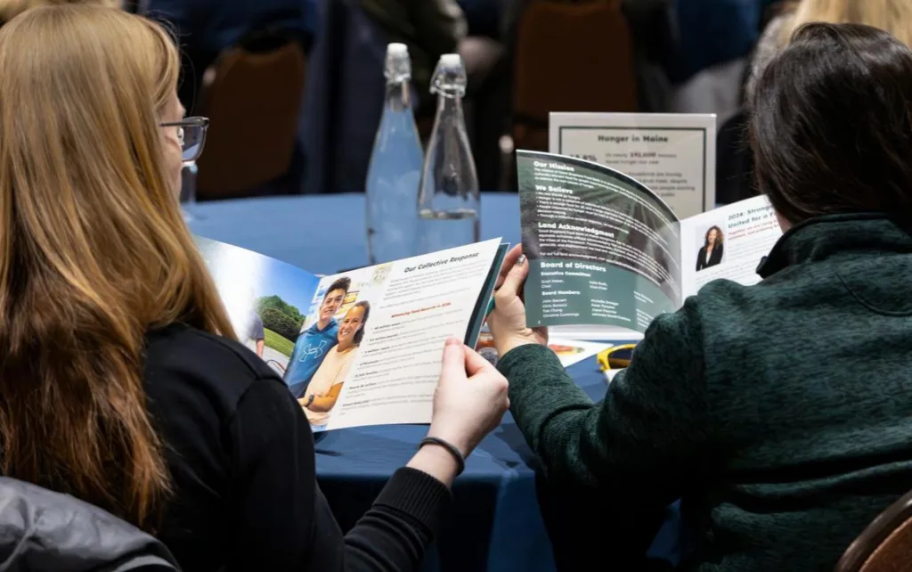 Two people sitting at a table read brochures at an event; water bottles and informational signs are on the table in front of them.