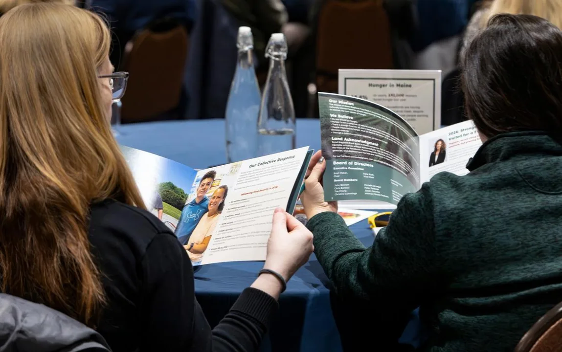 Two people sitting at a table read brochures at an event; water bottles and informational signs are on the table in front of them.