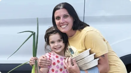 Hayley and her little girl, Maizey, are smiling and holding up some of the nutritious food they chose at Youth Full Maine, a partner of Good Shepherd Food Bank.
