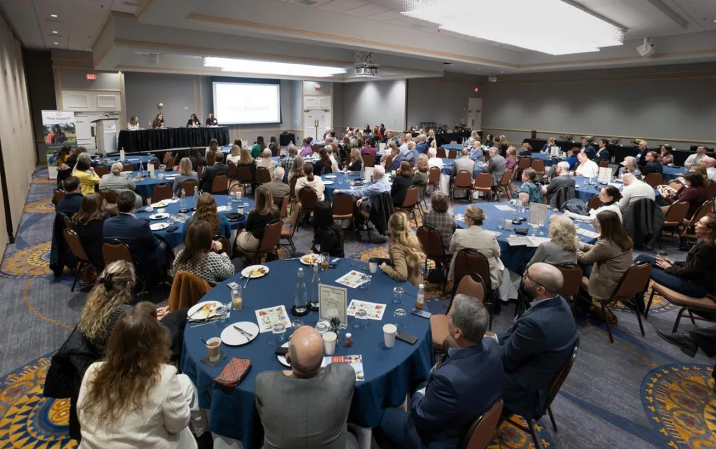 A large group of people sits at round tables in a conference room, facing a stage where panelists speak at a table with a presentation screen behind them.