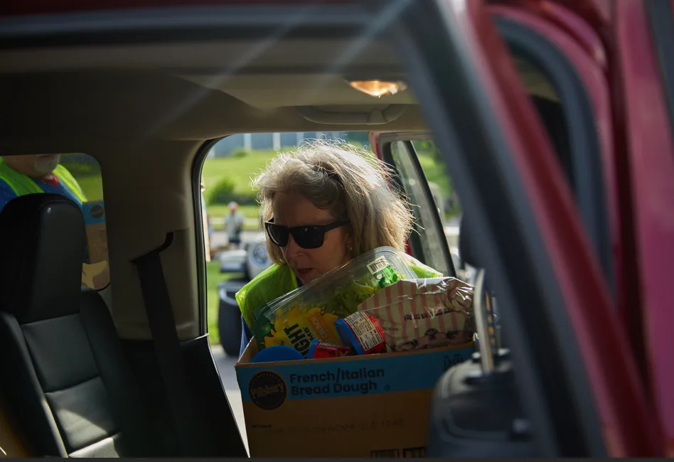 A woman wearing sunglasses and a safety vest loads a box of groceries into a car through an open door.