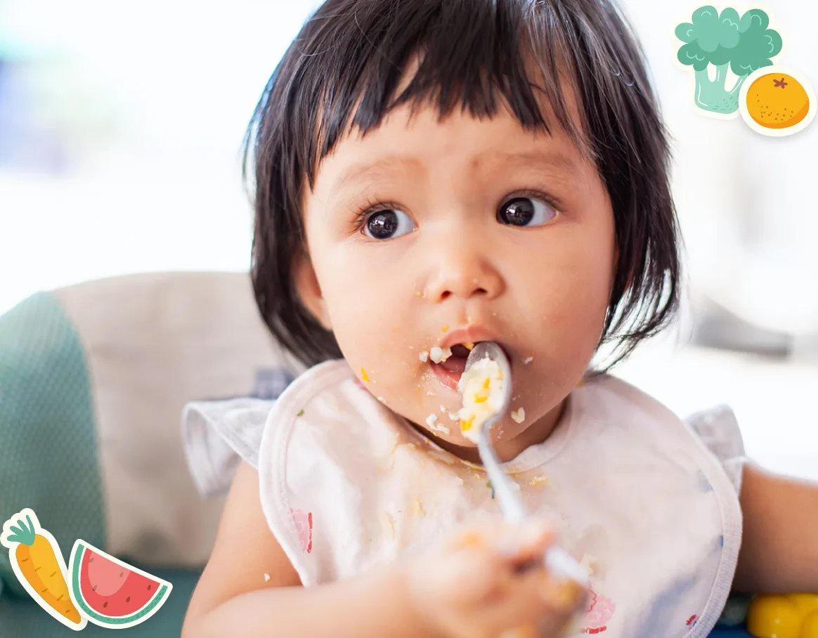 Give Now A toddler with short dark hair and a bib eats food with a spoon, with food around their mouth. Cartoon fruit and vegetable stickers decorate the image corners.
