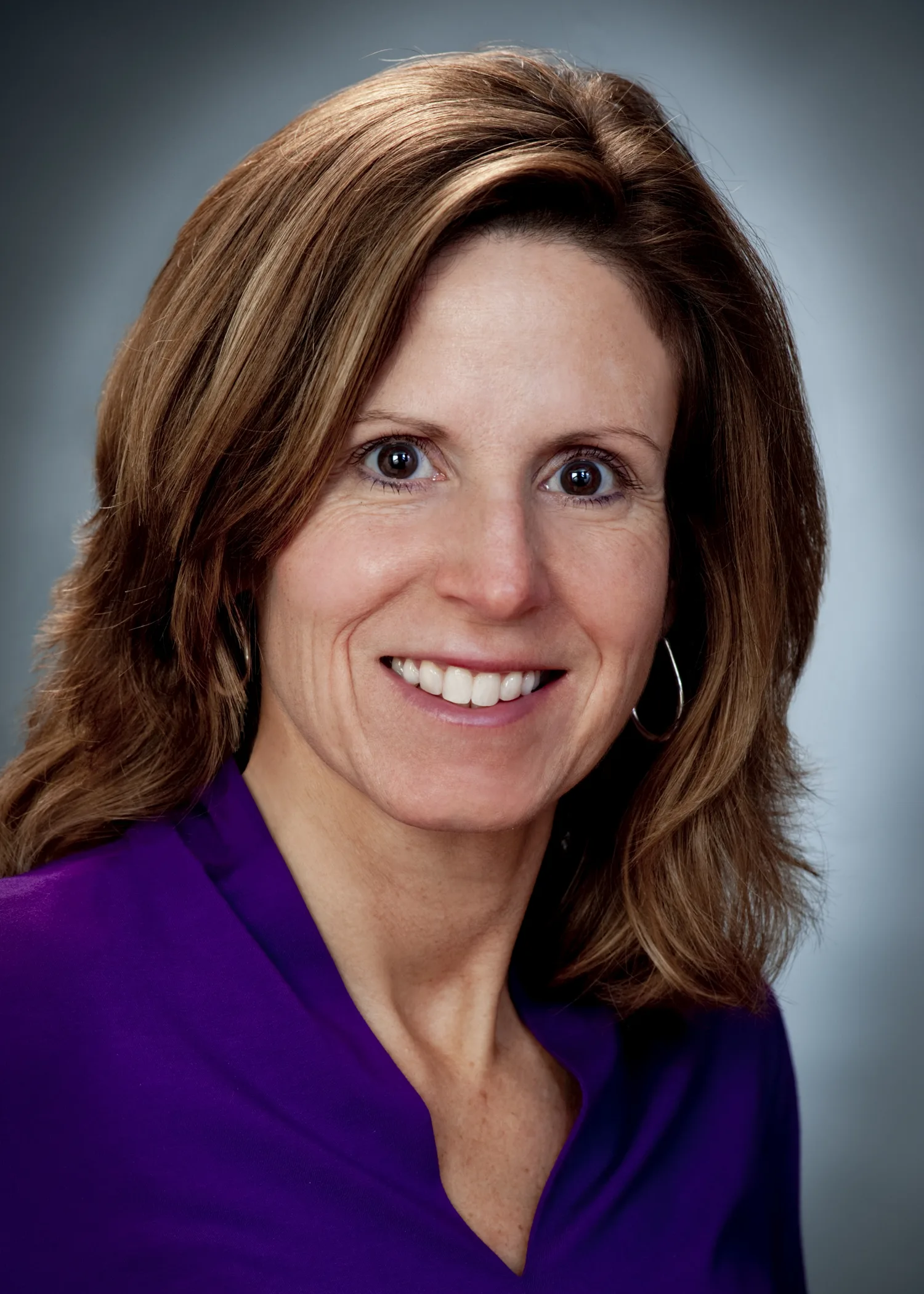 A woman with shoulder-length brown hair, wearing a purple blouse and hoop earrings, smiles at the camera against a gray background.