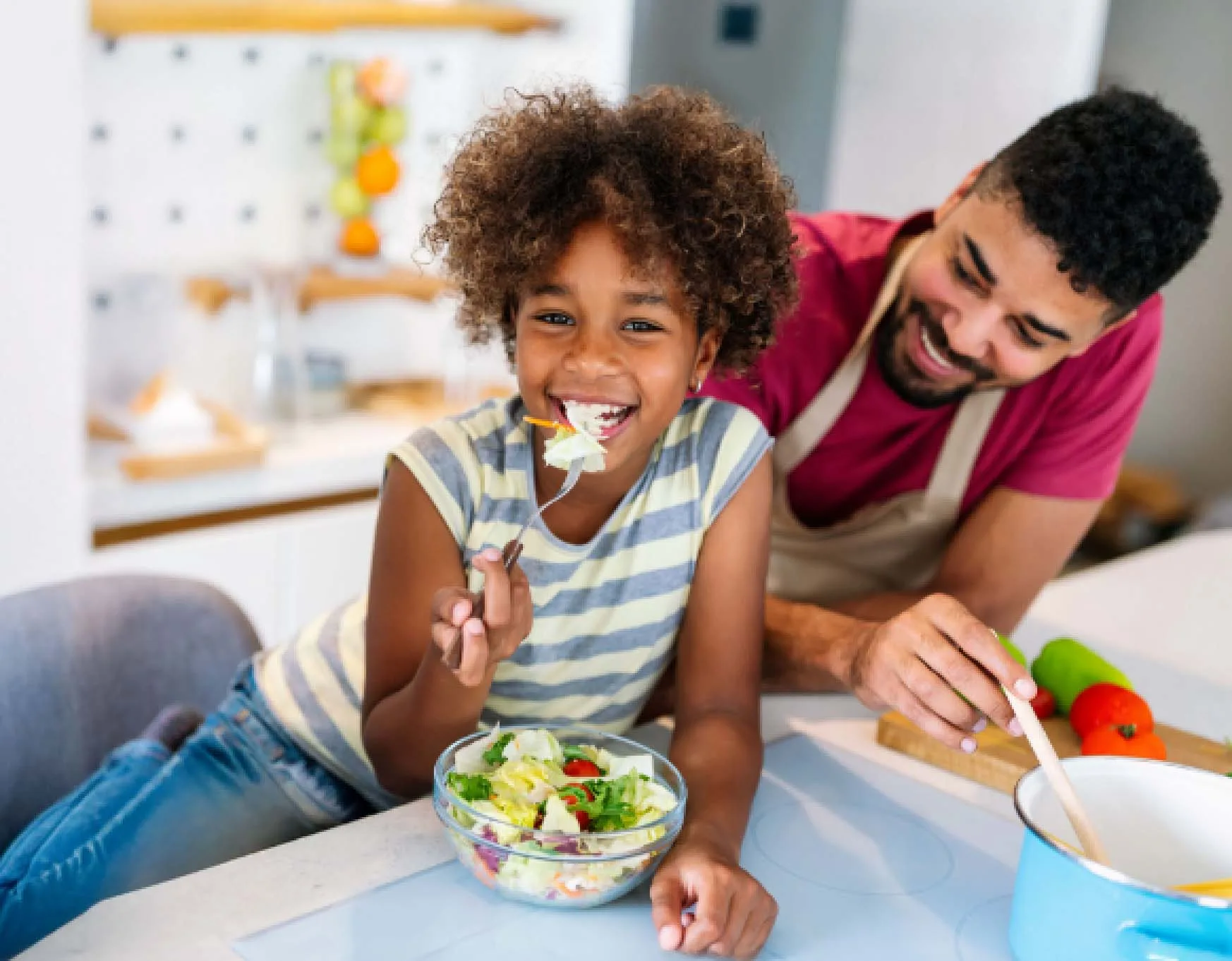 Give Now A smiling young girl eats salad at a kitchen counter while a man in an apron smiles beside her, preparing food.