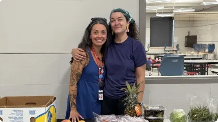 Tara and Courtney stand together behind tables loaded with food, ready for the next distribution.