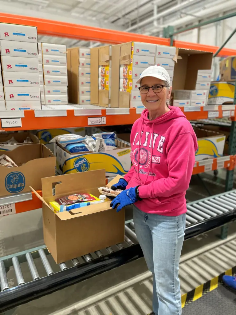 A smiling person in a pink hoodie and white cap packs food items into boxes on a roller conveyor in a warehouse.