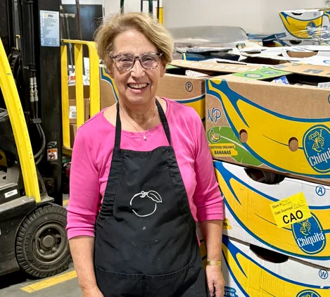 Smiling woman in a pink top and black apron stands beside stacked boxes of Chiquita bananas in a warehouse.