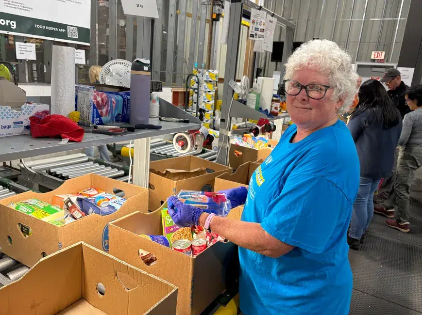 An older woman wearing a blue shirt and gloves sorts canned goods into boxes at a food bank with others working in the background.