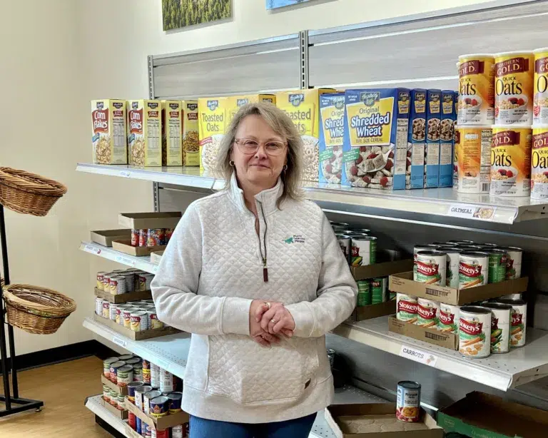 A woman in a light sweater stands in front of shelves stocked with cereal boxes and canned goods in a grocery store or food pantry.