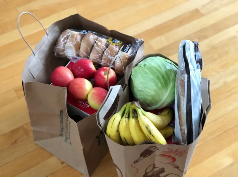 Two paper grocery bags on a wooden floor, filled with apples, bananas, cabbage, hot dog buns, and other assorted groceries.